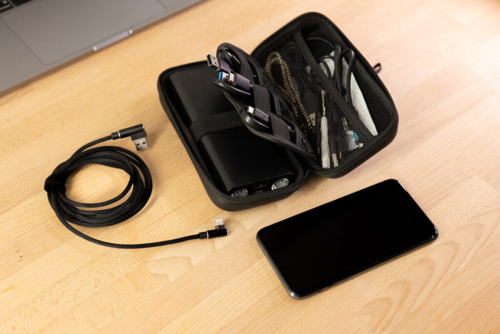 Cable organizer on a wooden table with a laptop, featuring black cables, a charger bag, and a power bank for travel