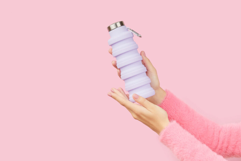 Hand holding and showing a reusable and collapsible lilac water bottle on a pink background