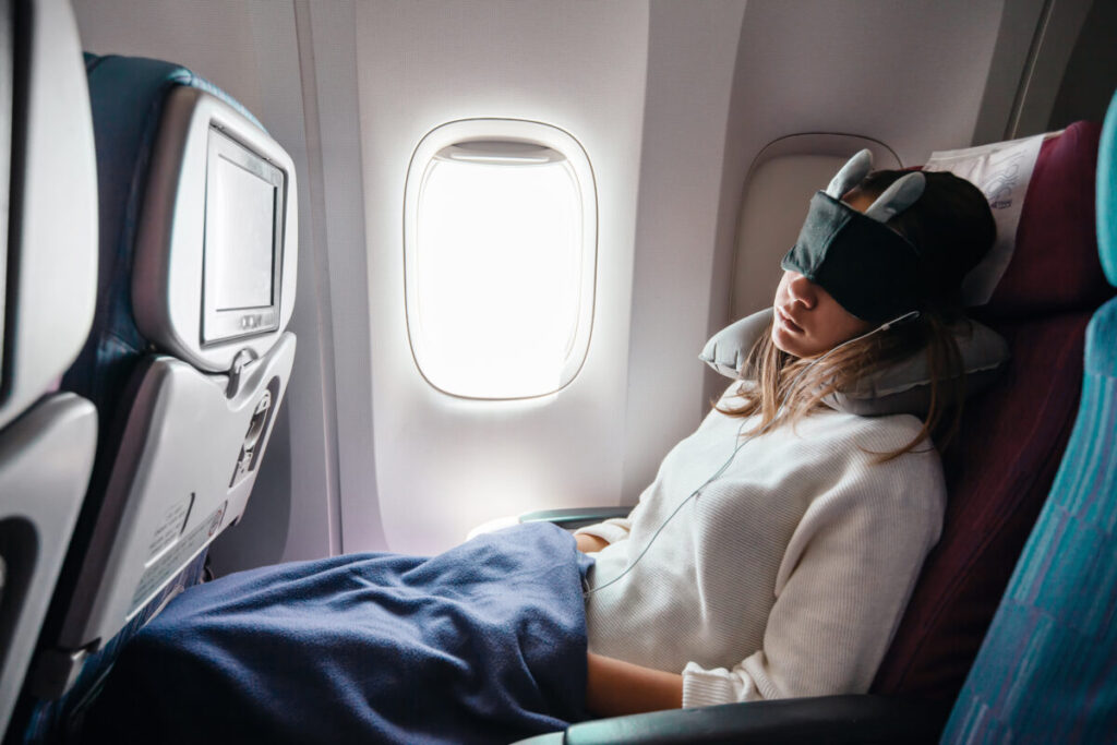 Female airplane passenger sleeping peacefully, wearing an eye mask and wrapped in a cozy travel blanket