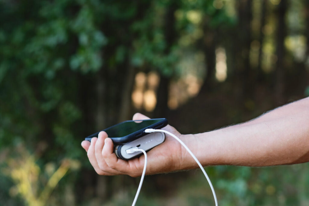 Man stands in nature, holding a smartphone and portable charger, with vibrant greenery behind him 